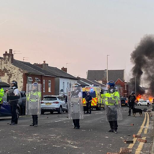 Smoke billows from a fire started by protesters as riot police stand guard after disturbances near the Southport Islamic Society Mosque in Southport, northwest England, on July 30, 2024, a day after a deadly child knife attack. Violent clashes broke out in the northern England town where a knife attack claimed the lives of three children, with around 100 protesters lighting fires and battling police. A 17-year-old male suspect from a nearby village arrested shortly after the incident remained in custody, police added, as they warned against speculating about his identity or details of the investigation. (Photo by Roland LLOYD PARRY / AFP)