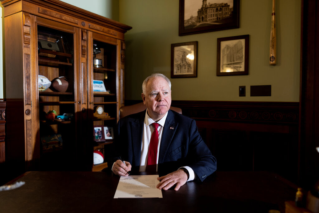 Gov. Tim Walz of Minnesota, wearing a blue suit and red tie, is sitting at a desk holding a pen above a sheet of paper.