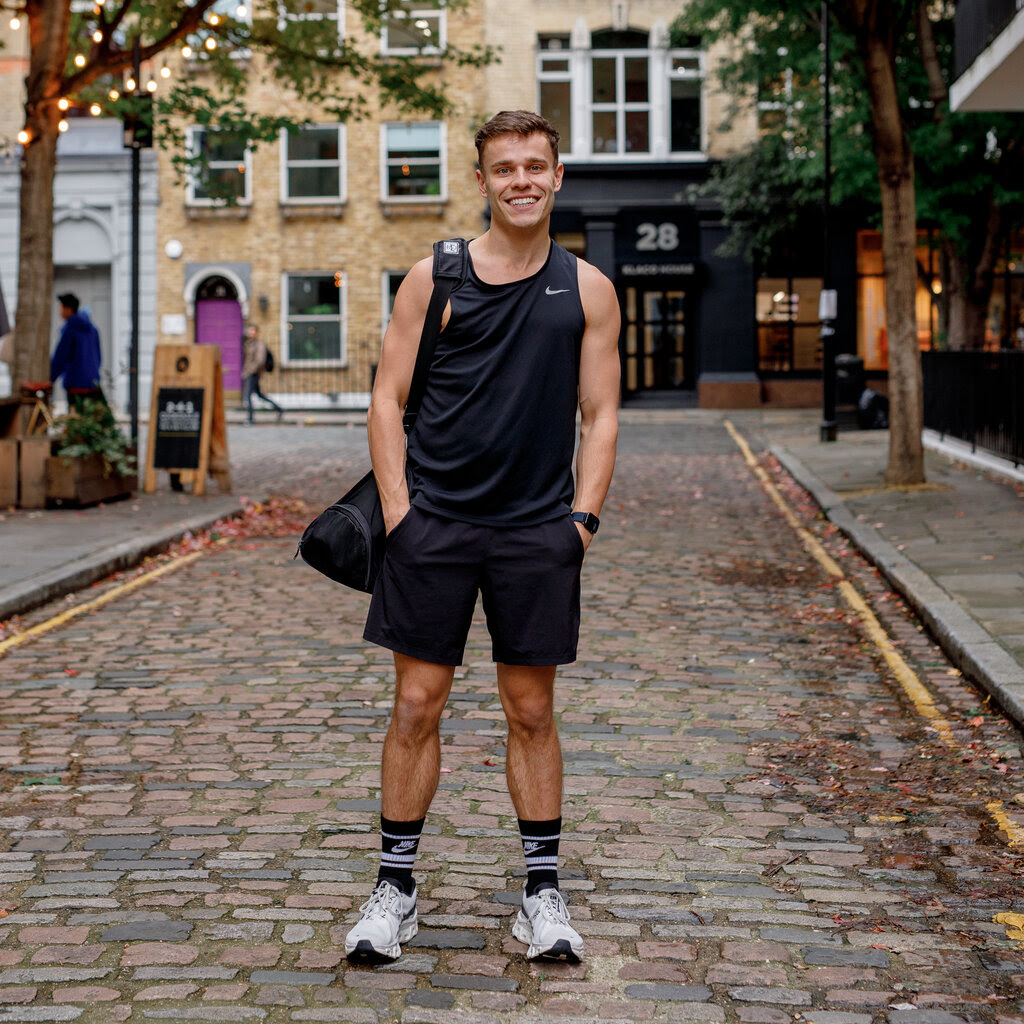 A man in workout clothes, holding a gym bag, smiles as he poses on a cobblestone street. 