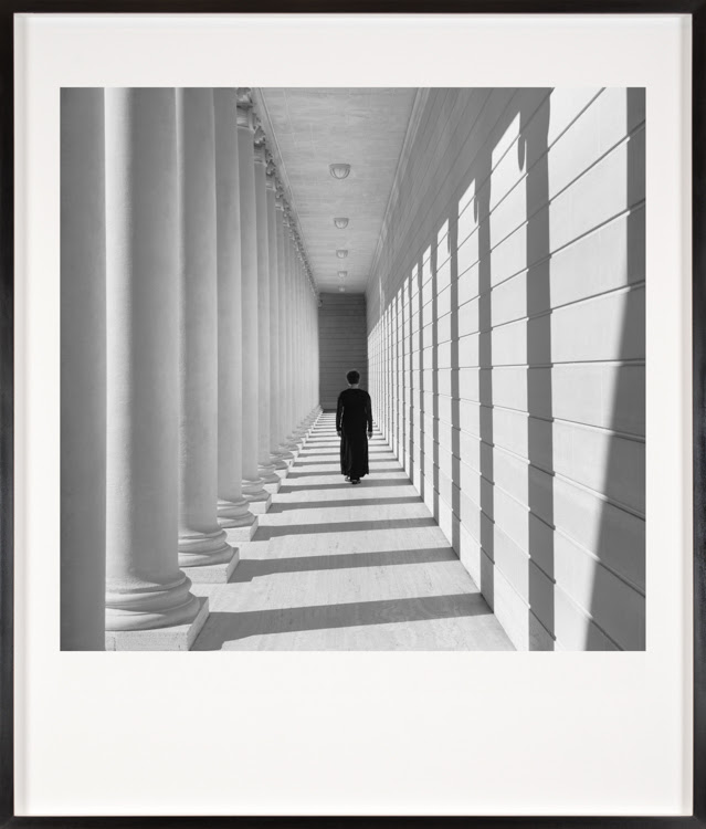 Color image of a framed black and white photograph of a woman in a black dress standing in the center of a hall lined with columns on one side and a wall on the other