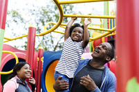 A family are at the playground, the child is hanging on a climbing frame, supported by her father