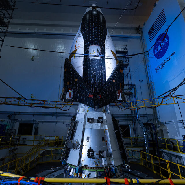 A space vehicle stands upright in a darkened facility with the NASA logo prominent on one wall.