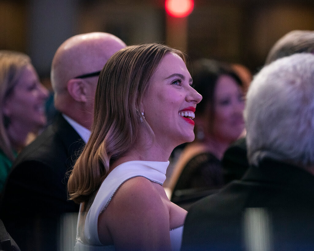 Scarlett Johansson, wearing a white dress, smiles among a room of other people.