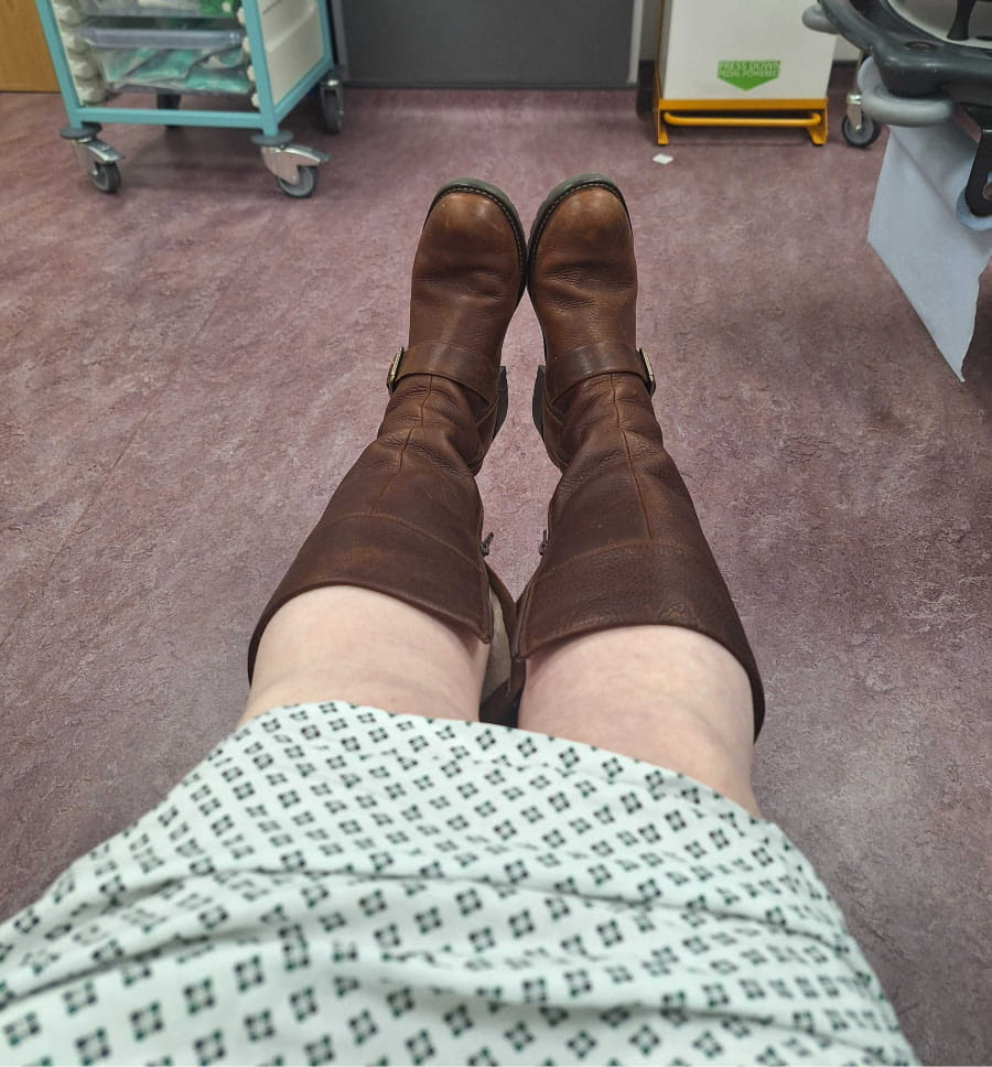 Photo of a woman's legs in knee-length brown boots, visible beneath a hospital gown, as she sits in a treatment room