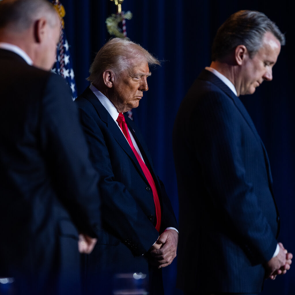 President Donald Trump and others in suits stand and bow their heads.