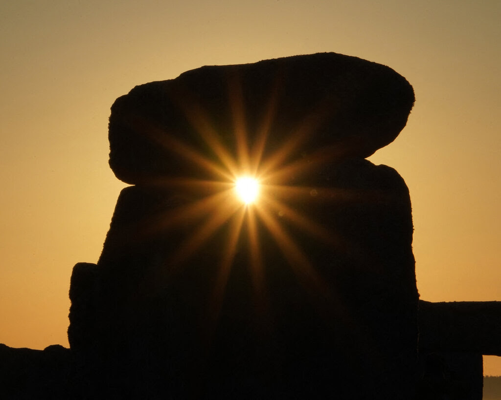 Sunlight flares through a hole in the silhouetted rocks of Stonehenge, creating a perfect golden starburst pattern.