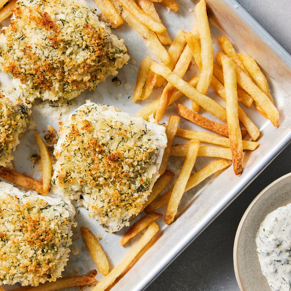 Four crumb-topped fish fillets are on a sheet pan with French fries; a small bowl of tartar sauce is nearby.