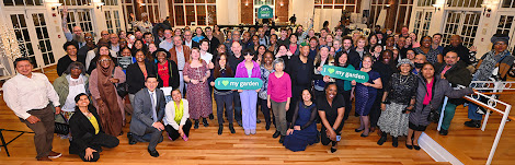 A large group of smiling people standing, sitting, and kneeling in a large bright room. Some people are holding garden recognition awards and signs saying I heart my garden.