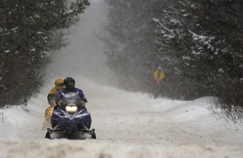 two people in helmets and snow gear ride on a snowmobile on a snow-covered trail surrounded by trees