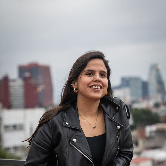 A woman wearing a black leather jacket with buildings in the background.