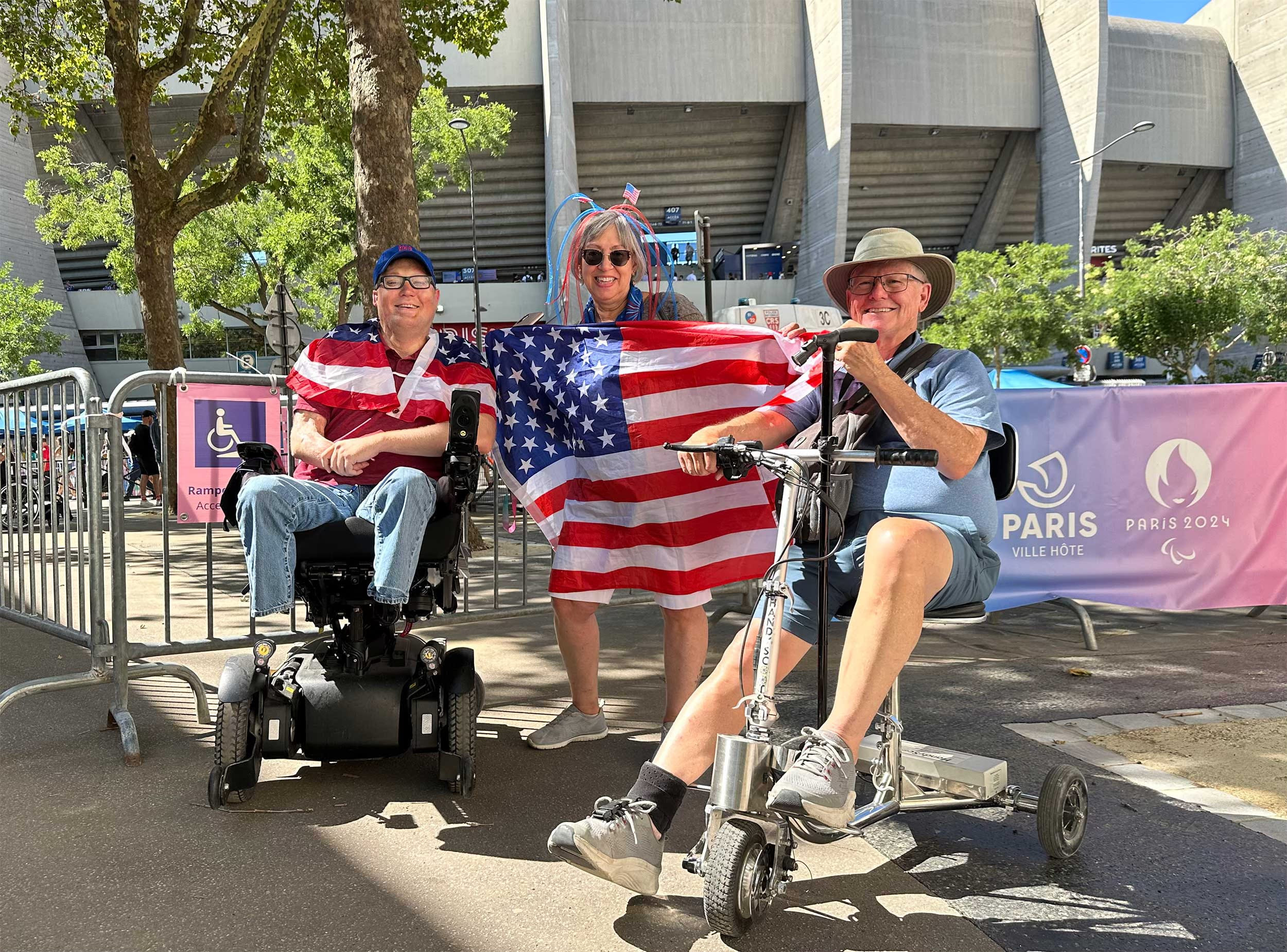 John seated in his wheelchair next to a man with a scooter and a woman holding a United States flag outside of a football stadium. John seated in his wheelchair next to a man with a scooter and a woman holding a United States flag outside of a football stadium.