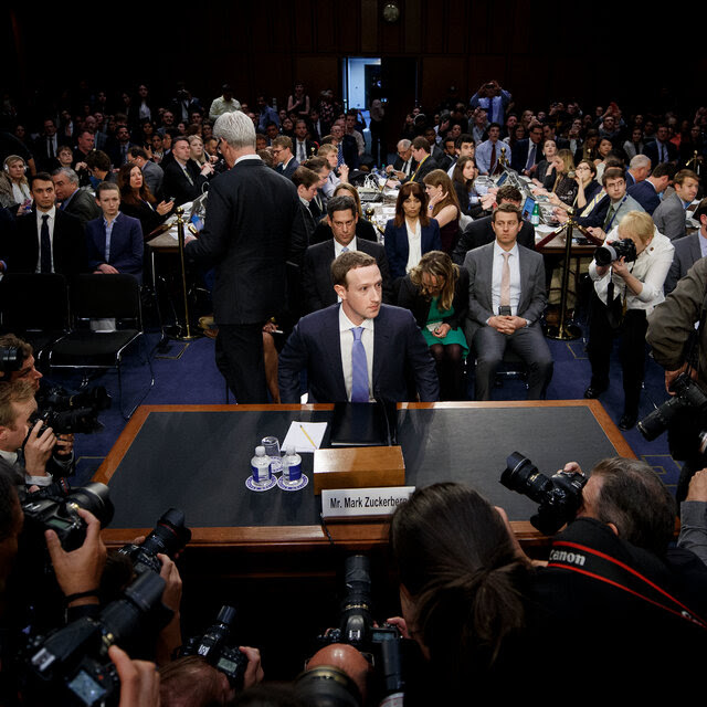 Mark Zuckerberg sits at a desk with an audience behind him and a crowd of photographers training their lenses on him in front of him. 