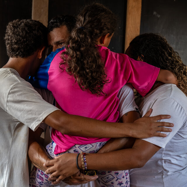 A group of four people, including a child in a pink shirt, embrace in a group hug.