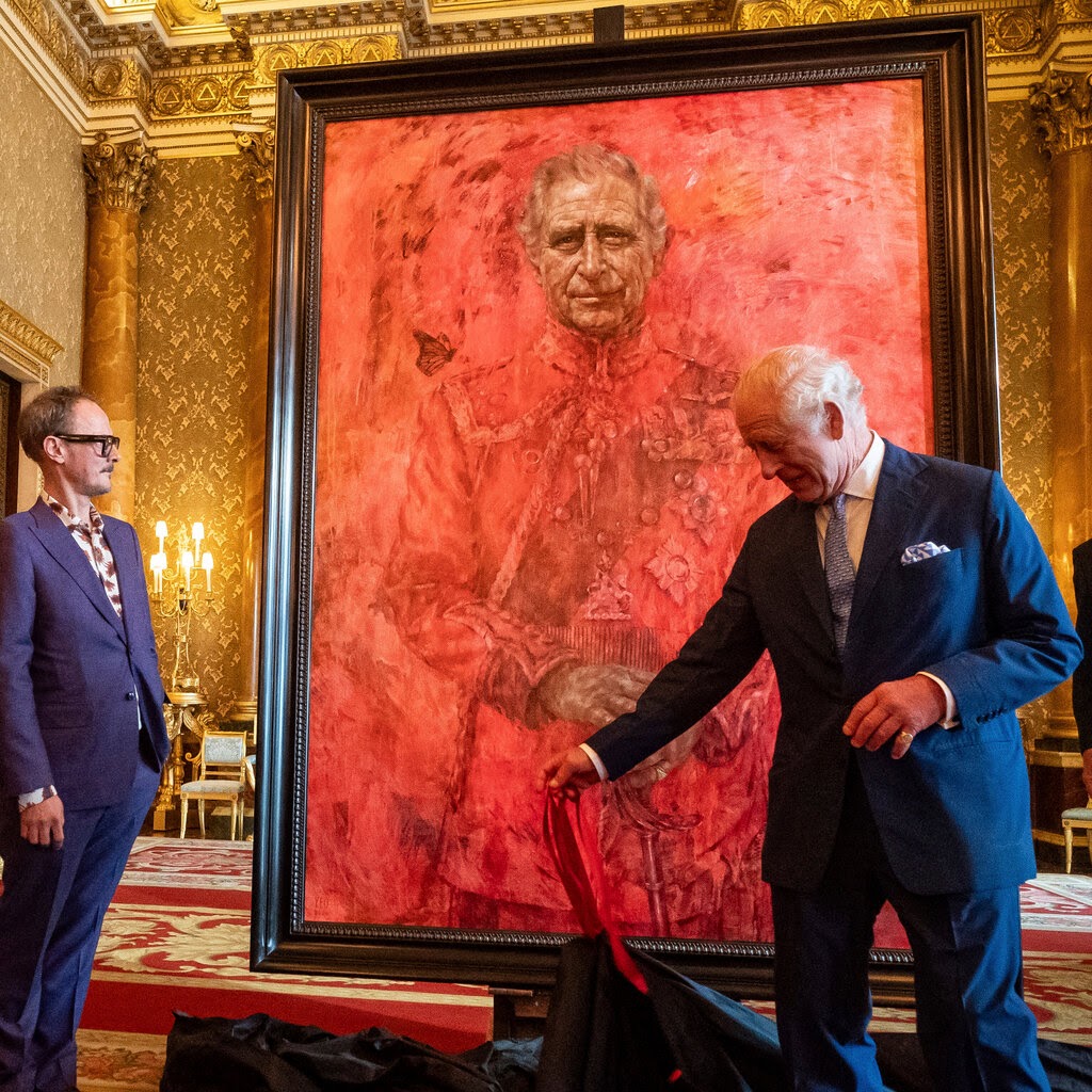 King Charles III stands in front of a large red portrait of himself. Another person, in a purple suit, stands nearby, looking at the painting. 