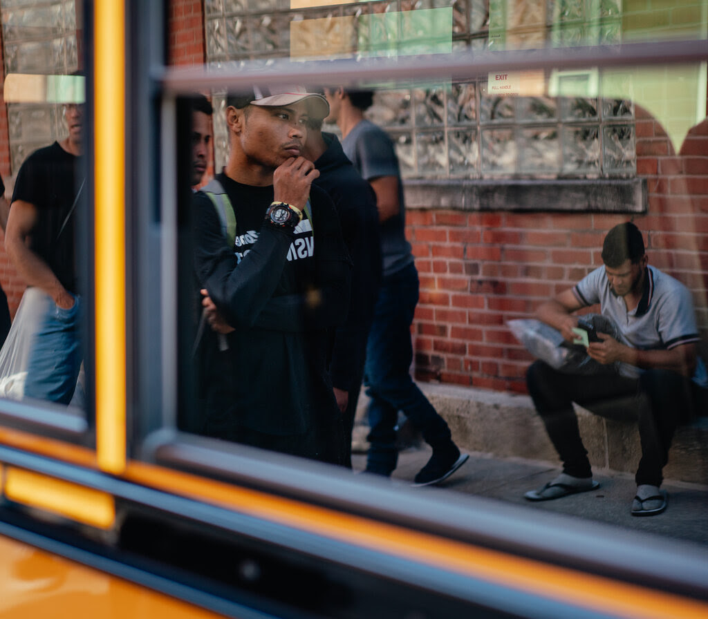 Men, seen in the reflection of a window, walk and sit along a sidewalk.