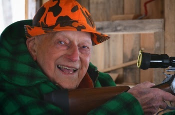 smiling, older man in orange and black camo hat and green plaid jacket holds a black rifle at window of a wood hunting blind