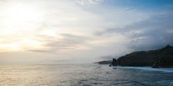 a large body of water, waves rolling toward the shoreline of a rising hill area, as the sun starts to rise through filmy white clouds