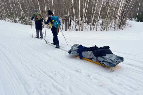 Two people cross-country skiing. One is pulling a sled filled with rolled up tarps.