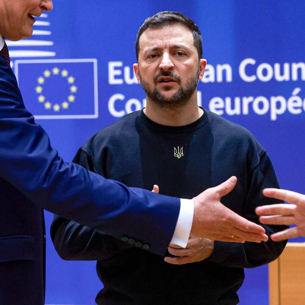 Volodymyr Zelensky standing in front of a blue background as a person in front of him reaches to shake another person’s hand.