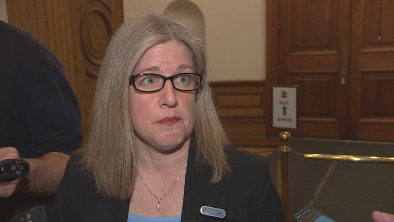 A woman with light hair and glasses stares as she is asked a question in a hallway.