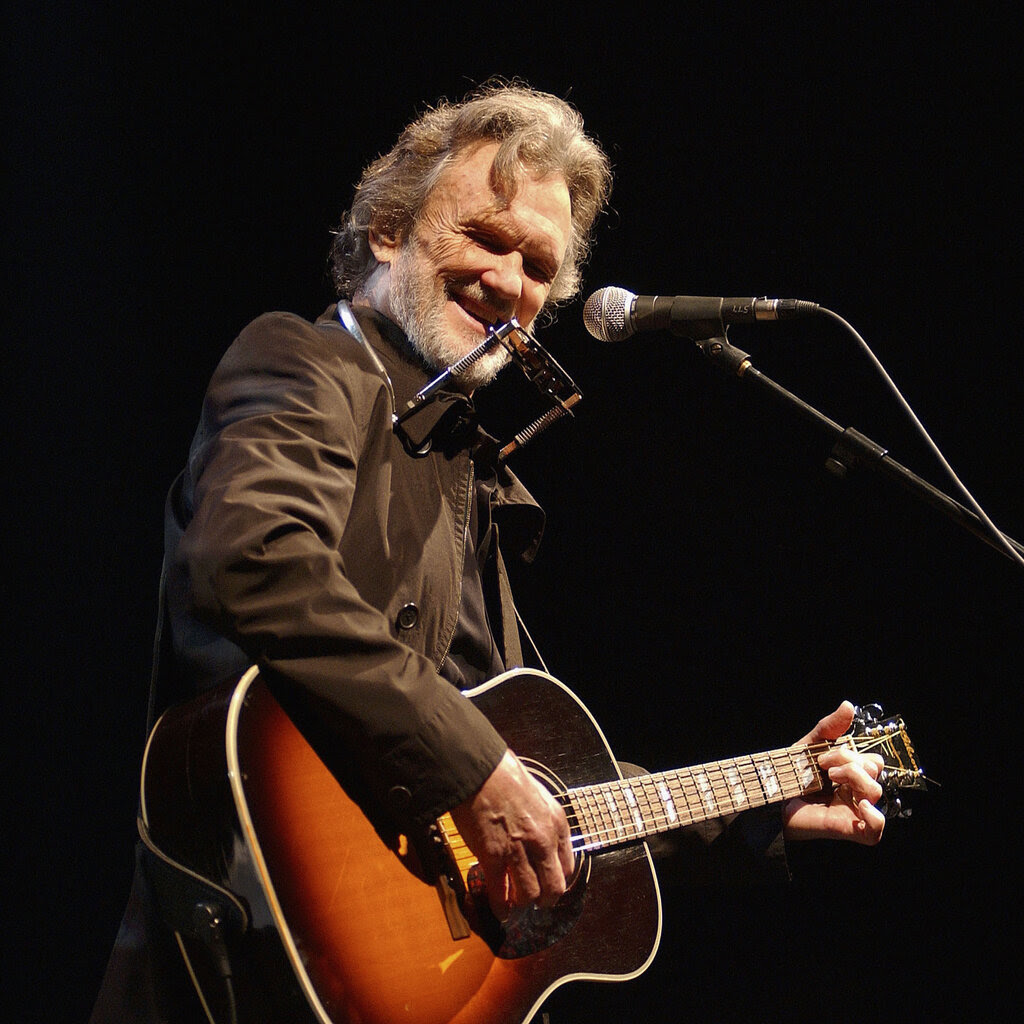 Kris Kristofferson in a brown jacket and a harmonica slung around his neck plays an acoustic guitar and grins, standing before a microphone.