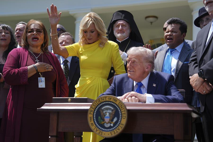 President Trump sits at a desk during a National Day of Prayer event. He is surrounded by many people.