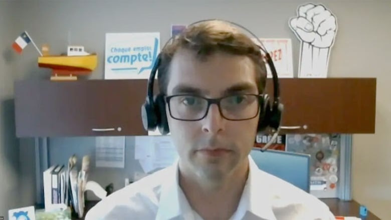 A man with glasses wears a headset and sits starting into a camera, with a shelf and desk against th wall behind him.