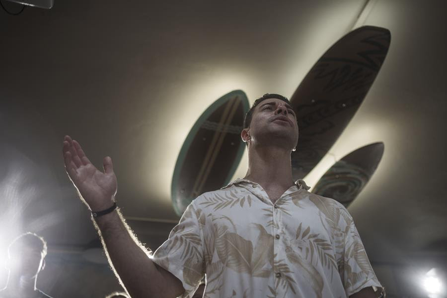 A man in a tropical shirt worships at church. There are three surfboards decorating the ceiling that are visible behind his head.