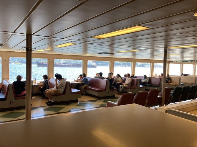 Several people sitting in booths along the windows inside the passenger cabin of a ferry