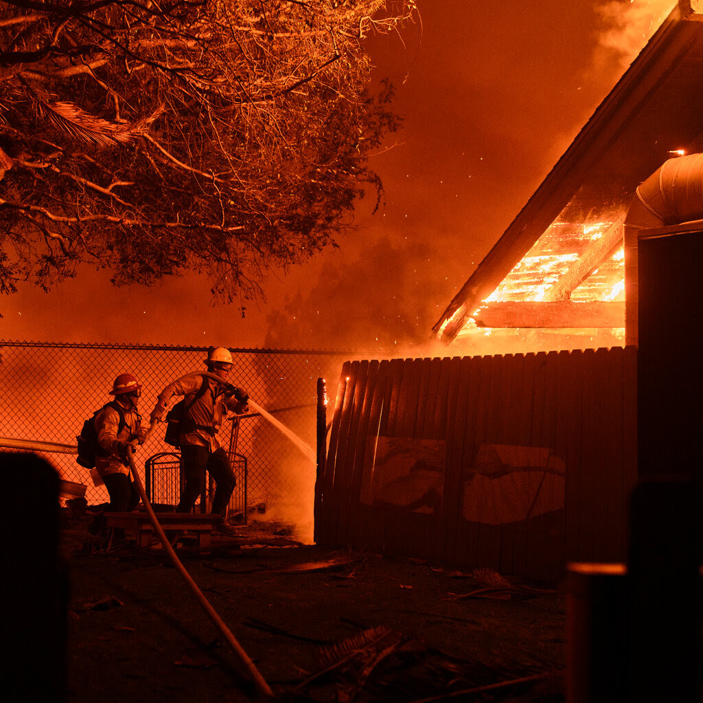 Two firefighters pulling a large hose that is shooting water toward a house on fire. 