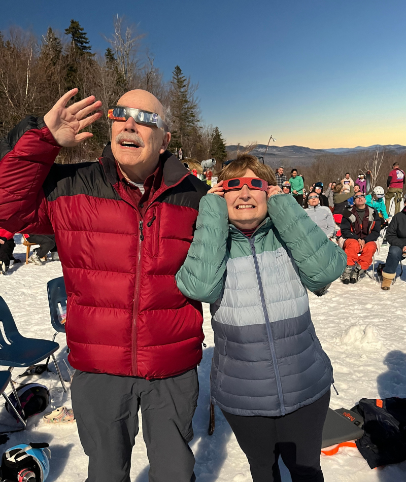 A man and woman stand in a crowd. Wearing jackets and eclipse glasses, they are watching the solar eclipse on April 8, 2024.