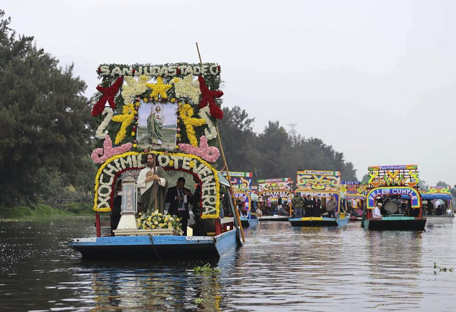 A procession of boats that are decorated with colorful flowers. The boat at the front of the procession has a glass case with a wooden figure holding a bone fragment of St. Jude.