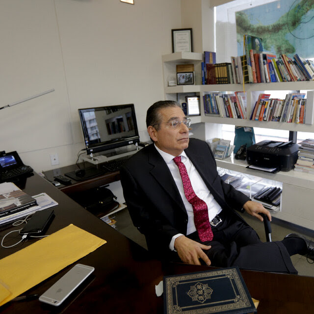 A man wearing a dark suit, white shirt and red tie sits behind a desk with his legs crossed, and a bookshelf behind him.