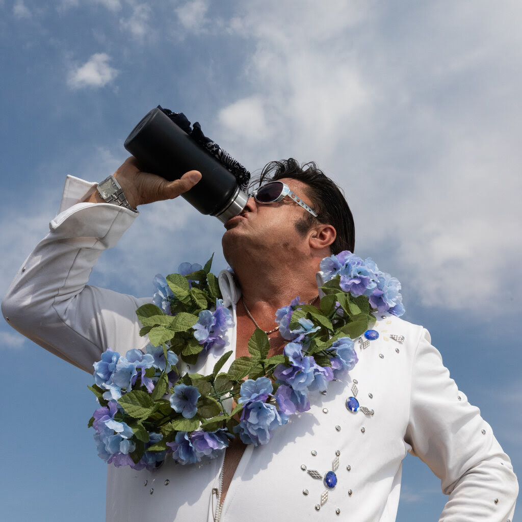 An Elvis impersonator, wearing sunglasses with a white sequined jacket and a lei, drinks deeply from a black metal water bottle.