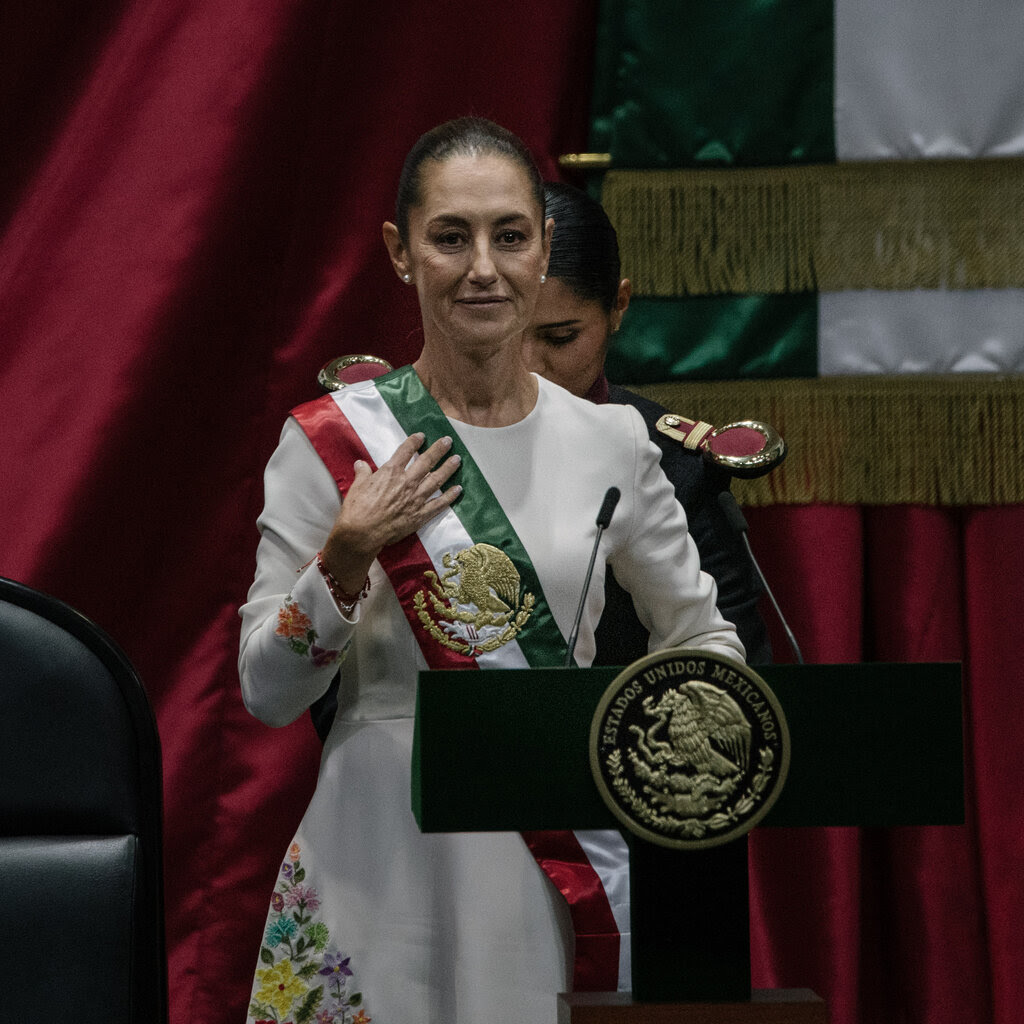 Claudia Sheinbaum, wearing a red, white and green sash, stands behind a podium. 