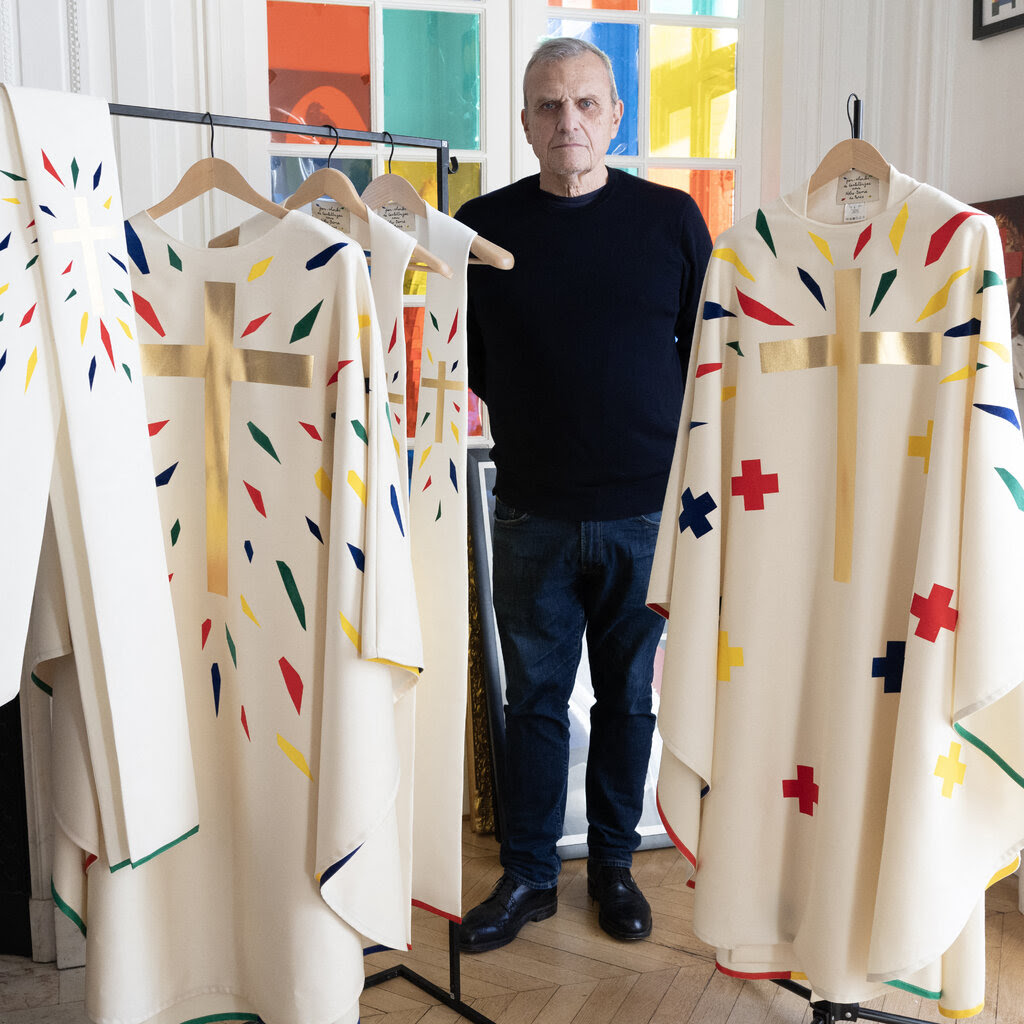 A man dressed in black, poses next to a rack hung with the poncho-like chasubles he designed. Patches of red, green, blue and yellow surround a large cross on the front of the white garments.