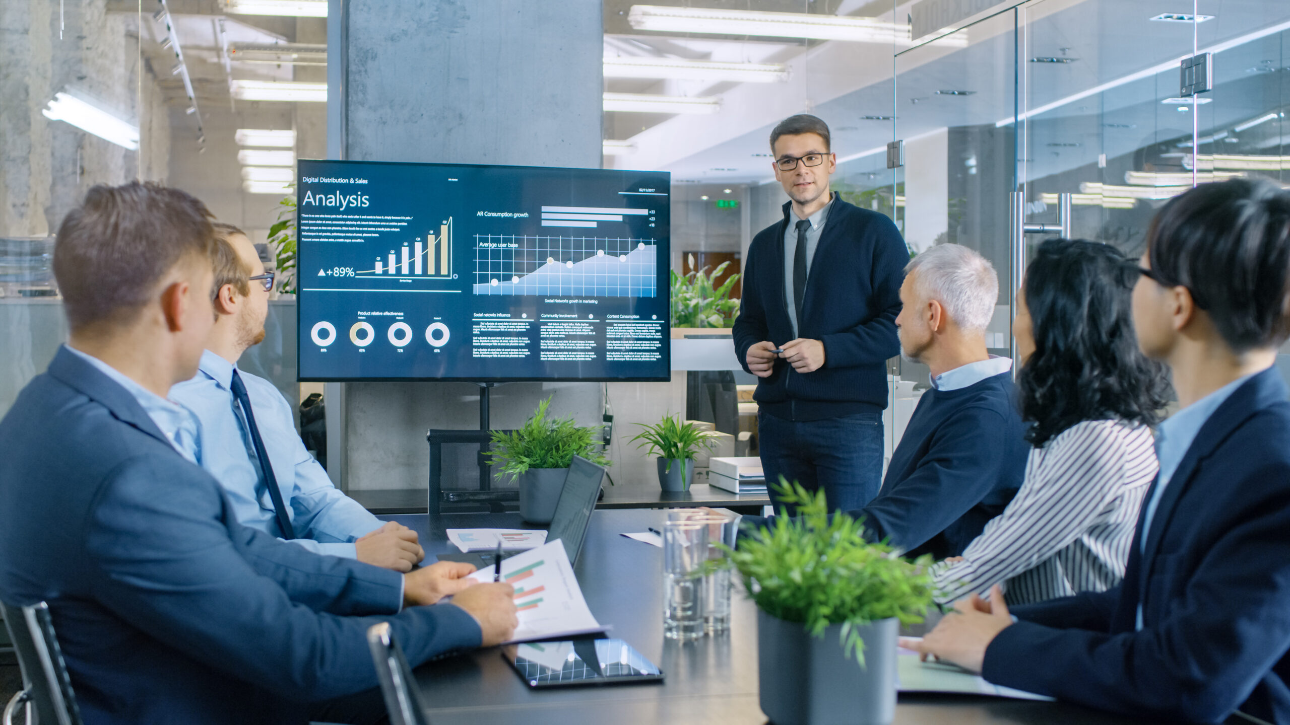 A diverse group of professionals gathered around a conference table in a sleek, modern boardroom. Charts and graphs are displayed on a large screen at the front of the room, while individuals engage in lively discussion, gesturing towards the data projections.