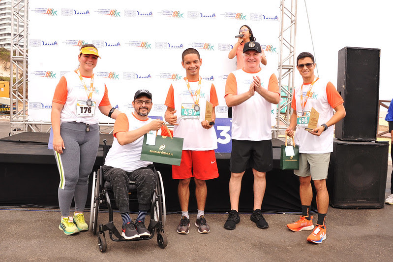 A imagem mostra quatro indivíduos lado a lado, cada um com uma medalha no pescoço e segurando o que parece ser um número de corrida na frente deles, dentre elas o presidente do TJMA, desembargador Froz Sobrinho, e ganhadores da corrida. Eles estão em frente a um fundo com vários logotipos da corrida, dado as medalhas e os números .