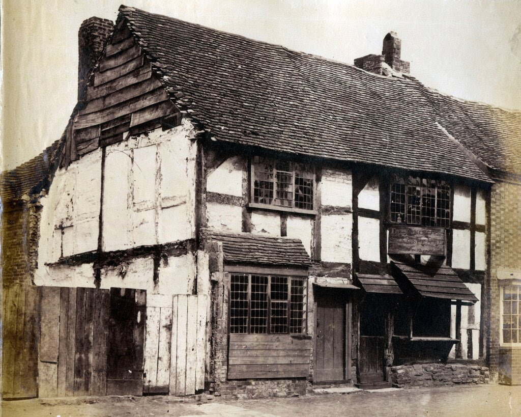 A black-and-white photo of a dilapidated three-story Tudor-style house with a steep shingled roof, before it was renovated in the late 1800s. The building is believed to be Shakespeare’s birthplace. The edges of the photo are yellowing.