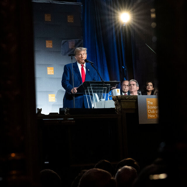 Donald Trump speaks into a microphone at a glass lectern onstage. 