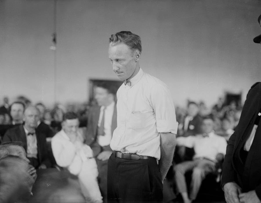 A man in a white shirt looks downcast as he stands among rows of seated people.