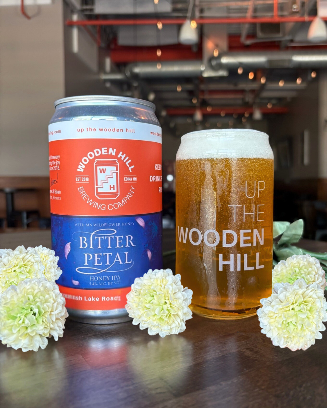 Picture of our new Honey IPA, Bitter Petal. Pictured: White and green flowers surrounding can and glass of Bitter Petal.