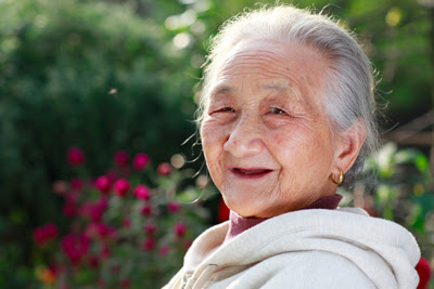 An older adult woman, with a toothless smile, is outside in the garden.