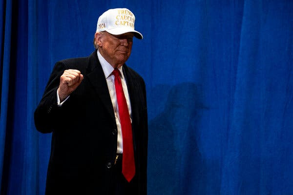 Donald Trump stands on a stage pumping his right fist. He’s wearing a dark suit, white shirt, red tie and a white baseball cap that says Trump Caucus Captain.