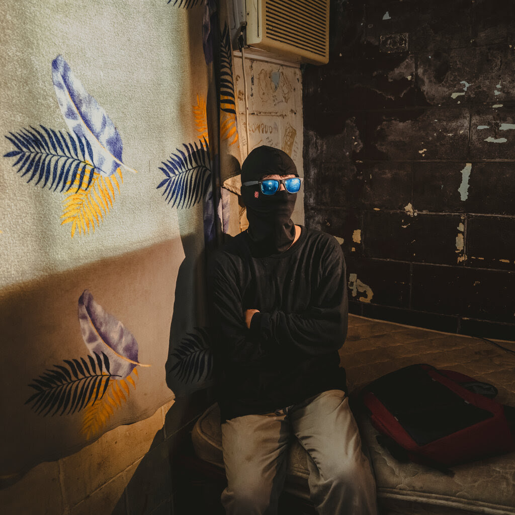 A person sits on a dirty mattress wearing a full ski mask and sunglasses to mask his identity. 