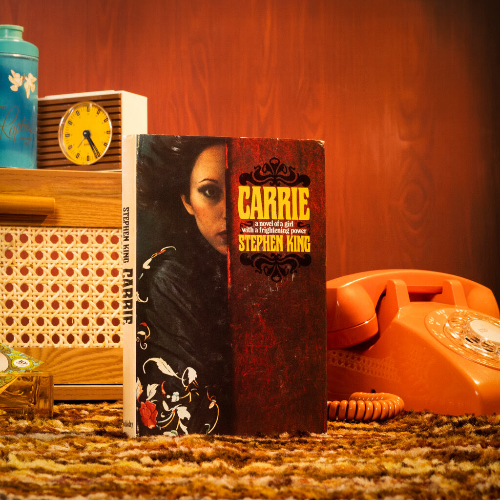 A still-life photo of a hardcover edition of “Carrie” on a brown shag carpet, next to an orange rotary-dial telephone and a section of chair caning with an analog clock balanced on top. The wall behind them is paneled wood.