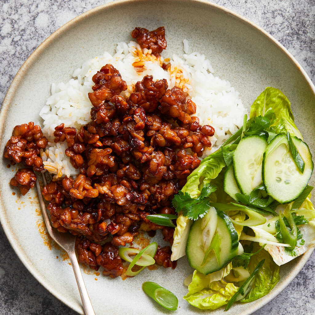 A spicy tempeh mixture served over ride with a side salad on a white plate.