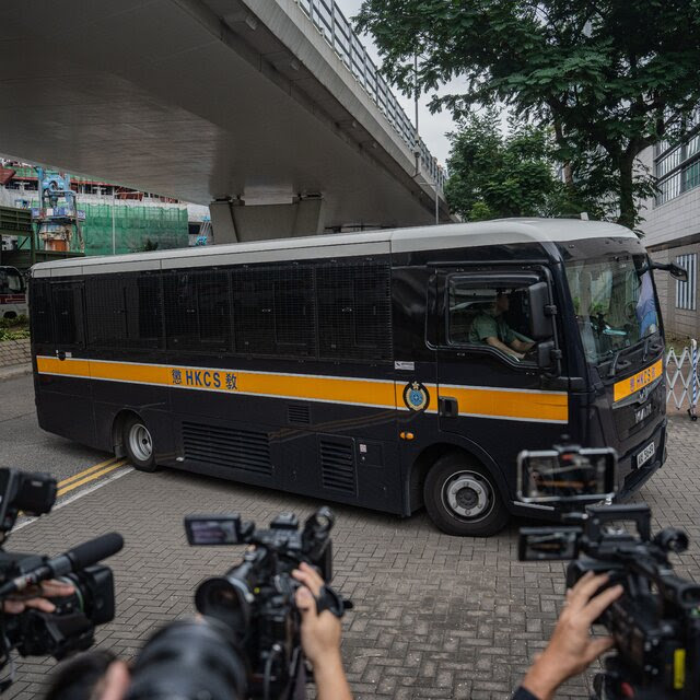 A black bus labeled “HKCS” turns off a road, toward a building. People with video cameras are in the foreground, filming. 