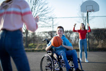 a young boy in a wheel chair is holding a basketball in an outdoor court, he is about to pass it to one of his teammates