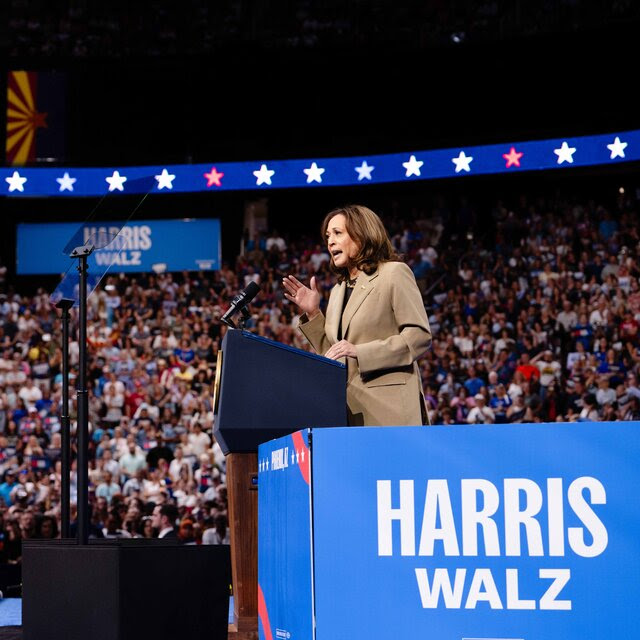 Kamala Harris, shown in profile, speaks from behind a lectern in a large arena filled with people. Several blue-and-white Harris-Walz campaign signs are displayed in the arena.